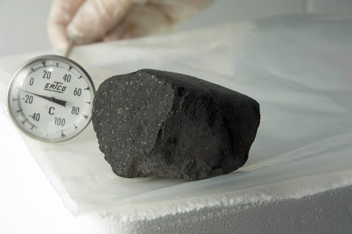 Meteorit vom Tagish Lake Schwarzer Stein mit kleinen weißen Einschlüssen auf einem Labortisch. Daneben wird ein Thermometer gehalten.