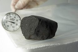 Schwarzer Stein mit kleinen weißen Einschlüssen auf einem Labortisch. Daneben wird ein Thermometer gehalten.