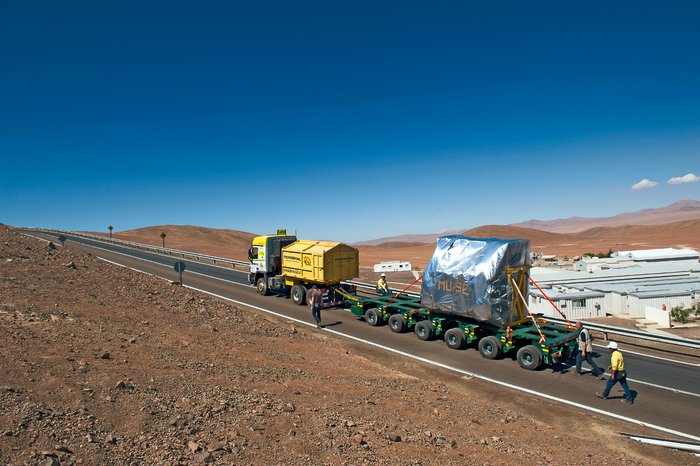 Auf einer Straße, die bergaufwärts in der steinigen Atacamawüste führt, zieht ein LKW das tonnenschwere MUSE-Instrument auf einem Anhänger. Neben und hinter dem Lastzug laufen einige Menschen mit Schutzhelmen.
