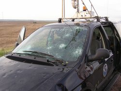 Foto. Beschädigtes Auto. Die Windschutzscheibe ist an mehreren Stellen sternförmig zersplittert. Die Motorhaube hat viele Dellen. Auf dem Dach des Autos sind technische Aufbauten zu erkennen.