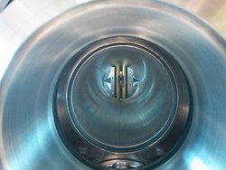 Blick in die Vakuumkammer, ein zylindrischer Hohlkörper aus Metall. Hinten im Bild eine kreisförmige Öffnung, in der senkrechte Goldbänder verlaufen. Zwischen den Bändern befindet sich ein quadratisches Loch, das den Durchmesser des Molekülstrahls begrenzt.