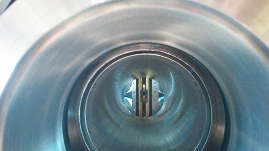 Blick in die Vakuumkammer, ein zylindrischer Hohlkörper aus Metall. Hinten im Bild eine kreisförmige Öffnung, in der senkrechte Goldbänder verlaufen, dazwischen ein quadratisches Loch.