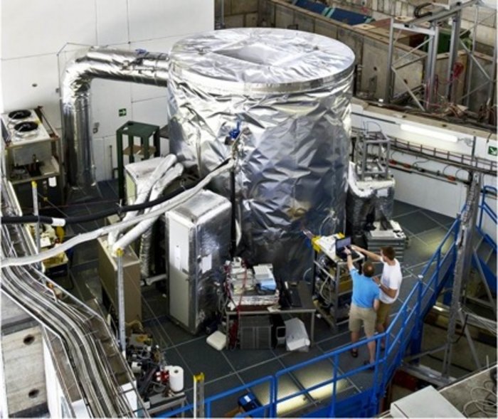Foto. In einer wissenschaftlichen Versuchshalle stehen technische Geräte. Im Zentrum befindet sich ein meterhoher metallischer Zylinder, der an Schläuche und kleinere Instrumente angeschlossen ist.