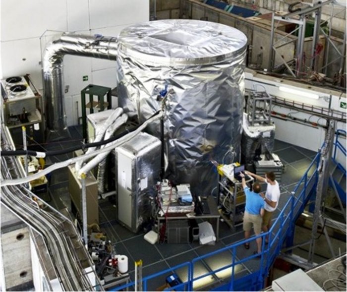 CLOUD-Versuchskammer Foto. In einer wissenschaftlichen Versuchshalle stehen technische Geräte. Im Zentrum befindet sich ein meterhoher metallischer Zylinder, der an Schläuche und kleinere Instrumente angeschlossen ist.