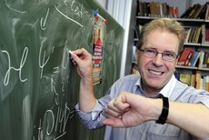 Foto von Peter Hänggi, der eine Brille trägt und gerade lächelnd mit einem Stück Kreide eine physikalische Formel an die Tafel schreibt