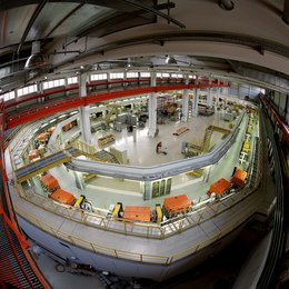 Weitwinkel-Aufnahme einer großen Halle, in der eine langgezogene Maschine in einem ovalen Aufbau steht. Entlag der Maschine sind zahlreiche Gerätschaften.