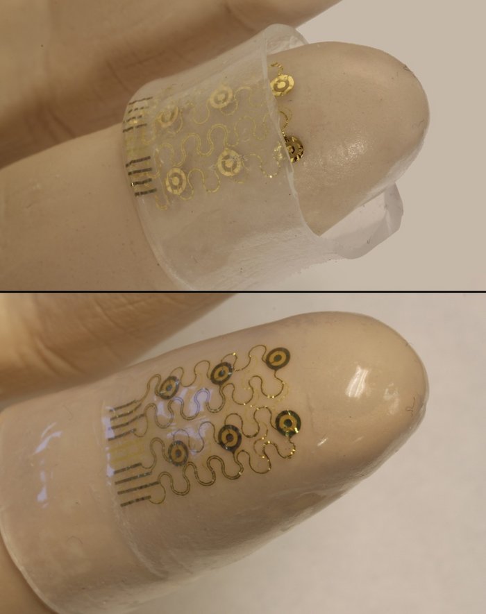 Tastempfindliche Fingerlinge Das Bild zeigt eine künstliche Hand aus Kunststoff. Über einer Fingerkuppe sitzt eine Haube aus einem durchsichtigen Material. Unter der Haube sind flache, goldene Elektroden zu sehen, das durchsichtige Material wird eng um Elektroden und Finger gelegt und passt sich deren Form an.