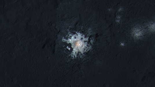 Eine dunkle Planetenoberfläche, im Zentrum des Bilds eine helle, von Furchen durchzogene, runde Region, die am Rand unregelmäßig ins Gestein ausläuft.