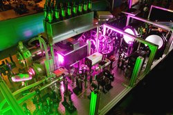 Auf einem Labortisch steht ein Laseraufbau mit Spiegeln, Linsen, Blenden und anderen optischen Geräten in grünes und violettes Licht gehüllt.