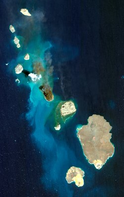 Von oben aufgenommene Inseln des Zubair-Archipels, die wie zackige Flecken auf dem Meer liegen, links oben im Bild eine kleine helle Rauchwolke.