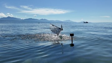 Ein vogelähnlicher Roboter mit zwei Flügeln ist knapp oberhalb der Wasseroberfläche eines Sees, das Wasser um ihn ist aufgewühlt, im Hintergrund sind Berge.