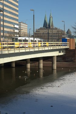 Eine Straßenbrücke in einer Stadt. Unter der Brücke ist offenes Wasser, daneben eine leich überschneite Eisschicht. Im offenen Wasser schwimmen einige Wasservögel.