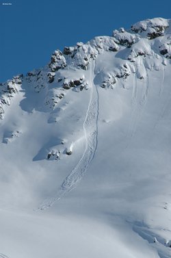 Foto: Schneebedeckter Berg, mit unberührter Schneedecke, dahinter wolkenloser Himmel. Im Schnee ist der Verlauf einer Lawine zu sehen, die oben im Berg an einem Punkt begann und sich nach unten hin immer weiter verbreiterte, wie ein Fächer, bis zu dem Ort, wo die Lawine als Schneehaufen zu liegen kam.