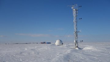 Das Bild zeigt eine schneebedeckte Landschaft mit einer kugelförmigen Messstation in der Mitte und einem Sendemast am rechten Bildrand. 