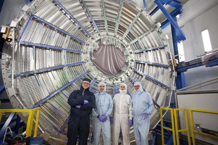 Das Bild zeigt den Spurdetektor des ALICE-Experiments von außen. Vor dem Detektor steht ein Team aus vier Wissenschaftlern.