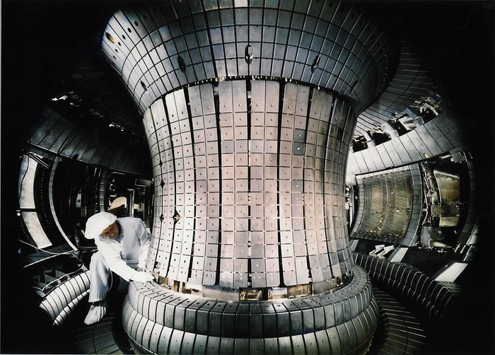 Plasmagefäß des Fusionsexperiments ASDEX Upgrade Foto: Techniker im Overall im Innenraum der Fusionsreaktors, im ringförmigen Tunnel rund um eine mehrere Meter hohe Mittelsäule. Diese mit glänzenden Kacheln verkleidet, die anderen Wände mit größeren Platten und Gitter-Verkleidungen für Vorrichtungen.