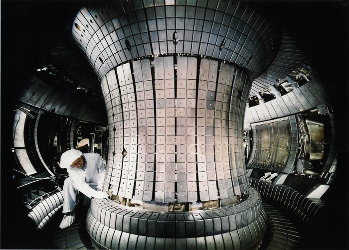 Foto: Techniker im Overall im Innenraum der Fusionsreaktors, im ringförmigen Tunnel rund um eine mehrere Meter hohe Mittelsäule. Diese mit glänzenden Kacheln verkleidet, die anderen Wände mit größeren Platten und Gitter-Verkleidungen für Vorrichtungen.