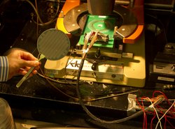 Wafer in Form eines Tischtennisschlägers mit einer Oberfläche aus kleinen goldenen Plättchen. Dahinter sind weitere elektronische Aufbauten zu sehen.