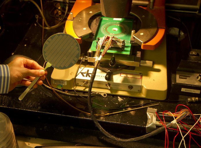 Prozessoren aus Kohlenstoffnanoröhrchen Wafer in Form eines Tischtennisschlägers mit einer Oberfläche aus kleinen goldenen Plättchen. Dahinter sind weitere elektronische Aufbauten zu sehen.