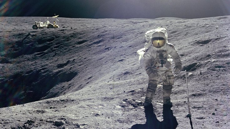 Astronaut steht am Rand eines Kraters auf dem Mond, im Hintergrund ist der Mondrover zu sehen.