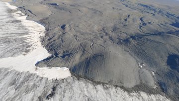 Foto einer Küstenregion: Am linken unteren Bildrand sind weiße Gletschermassen erkennbar, am oberen Rand das Meer