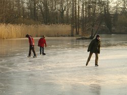 Drei Menschen auf einem zugefrorenen See. Sie bewegen sich auf Schlittschuhen. Es sind Spuren ihrer Fahrten zu erkennen, an diesen Stellen ist das glatte Eis durch Furchen aufgerauht.