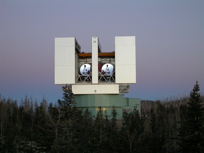 Das LBT auf dem Mount Graham in Arizona Großes, weißes, eckiges Gebäude oberhalb von Baumwipfeln. Das Dach ist geöffnet, im Inneren sieht man zwei große Spiegel.