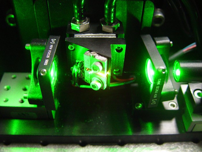 Grünes Pumplaserlicht in grünes Licht getauchtes optisches Experiment, Kabel und Schläuche, Blenden und Prismen, oben ein zylinderförmiger Behälter, in dem es rötlich leuchtet.