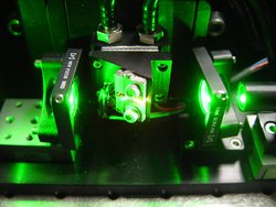 in grünes Licht getauchtes optisches Experiment, Kabel und Schläuche, Blenden und Prismen, oben ein zylinderförmiger Behälter, in dem es rötlich leuchtet.