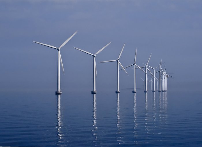Weiße Windräder eines Offshore Windparks spiegeln sich im blauen Wasser vor blauem Himmel.