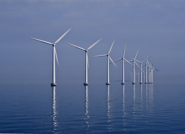 Offshore Windpark Weiße Windräder eines Offshore Windparks spiegeln sich im blauen Wasser vor blauem Himmel.