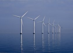 Weiße Windräder eines Offshore Windparks spiegeln sich im blauen Wasser vor blauem Himmel.