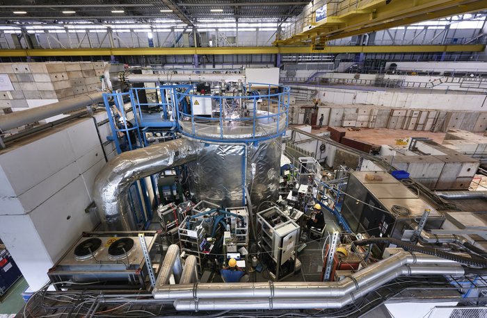 Das CLOUD-Experiment am CERN Blick in eine Halle mit verschiedenen Maschinen; in der Mitte steht ein silberner Kegel, um den mehrere Geräte gruppiert sind, an denen Menschen arbeiten