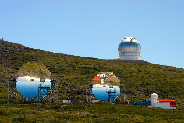 Die beiden MAGIC-Teleskope In leicht abschüssigem Gelände, mit Gras und Büschen bewachsen, steht neben dem ursprünglichen MAGIC-Teleskop ein baugleiches. In ihren Spiegeln zeigt sich die Landschaft von La Palma auf dem Kopf stehend. Rechts der beiden MAGIC-Teleskope steht ein Gebäude mit einer kleinen Kuppel und rotem Dach, das im rechten MAGIC-Spiegel erscheint. Im Hintergrund befindet sich am Horizont eine Teleskopkuppel in gleicher scheinbarer Größe wie die MAGIC-Spiegel, aber deutlich größerer Entfernung.
