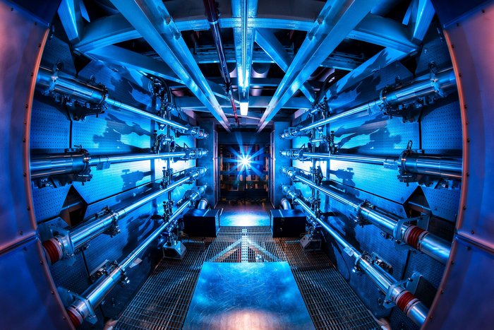 Das Lasersystem der National Ignition Facility Ein enger Gang mit Röhren an den Wänden, am Ende des Ganges leuchtet ein helles, blaues Licht.