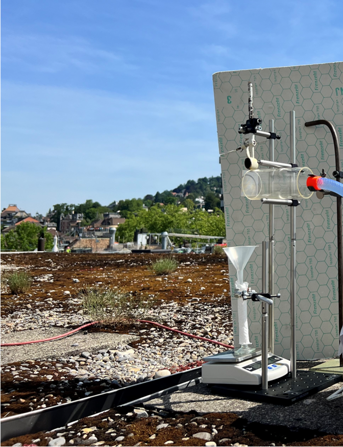 Versuchsanordnung unter freiem Himmel Versuchsaufbau auf einem Dach unter freiem Himmel: eine transparente Röhre ist an einem Metallständer befestigt, vor ihrer Öffnung befindet sich ein Drahtgeflecht; davor auf dem Boden steht eine Glasröhre auf einer Waage; in der Öffnung der Röhre steckt ein Trichter