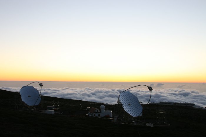 MAGIC-Teleskope im Sonnenuntergang Im Vordergrund der Berg, auf dem sich die Teleskope befinden. In der Mitte beide Teleskope, im Hintergrund eine darunter liegende dichte Wolkenschicht. Über der Wolkenschicht ist der Himmel klar und wolkenfrei.