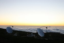 Im Vordergrund der Berg, auf dem sich die Teleskope befinden. In der Mitte beide Teleskope, im Hintergrund eine darunter liegende dichte Wolkenschicht. Über der Wolkenschicht ist der Himmel klar und wolkenfrei.