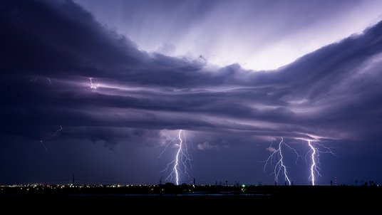 Das Bild zeigt Gewitterwolken und drei Blitze über einer Stadt. 