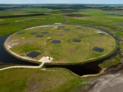 Eine weite grüne Wiese von oben betrachtet. Im Zentrum ein rundes Feld von einem Wassergraben umgeben, auf dem Antennen als dunkle Flecken zu erkennen sind.