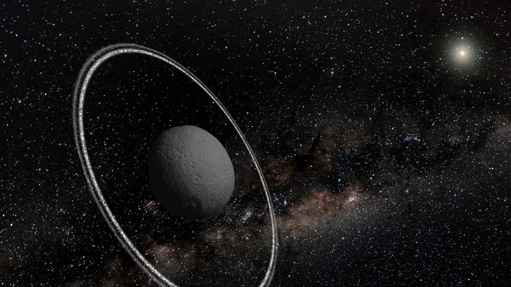 Asteroid vor Sternenhintergrund, hinten rechts die Sonne, der Asteroid ist von zwei dünnen Ringen umgeben.