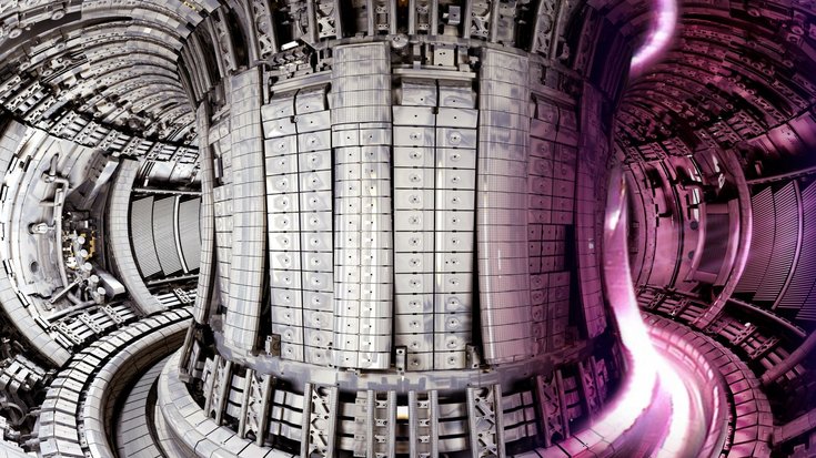 Blick in das Innere einer runden, metallenen Kammer. In der Mitte befindet sich eine große säulenartige Maschine; rechts davon ist ein rötlicher Lichtstrahl.
