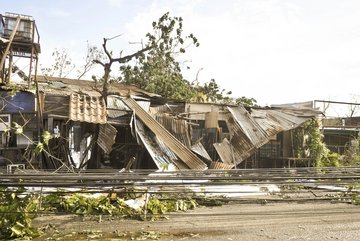 Durch einen Wirbelsturm zerstörte Gebäude mit eingedrückten Wellblechdächern, umgeknickten Bäumen und heruntergerissenen Leitungen.