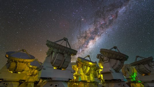 Mehrere, eng beieinander stehende Radioteleskope, im Hintergrund ein teil der Milchstraße.