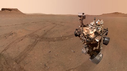 Marsrover Perseverance auf dem Mars. Sowohl Marsboden als auch der Himmel über dem Mars sind rötlich. Das Terrain ist flach und sandig. Im Hintergrund sind Hügel. Neben dem Rover liegen Bodenproben vom Mars in kleinen Tuben. 
