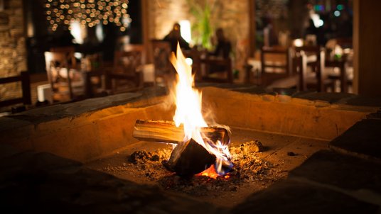 Ein brennendes Kaminfeuer steht im Mittelpunkt. Flammen leuchten warm über Holzscheiten und Glut. Dahinter verschwimmt ein gemütlicher Raum mit Holztischen, Stühlen und weichem, goldenem Licht, das eine ruhige, einladende Atmosphäre schafft.