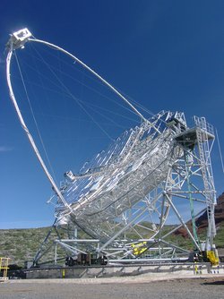 In strahlender Sonne ein silbrig glänzendes Gerüst in karger Landschaft. Die Hauptgitterstruktur ist erst von wenigen Metallplatten bedeckt.