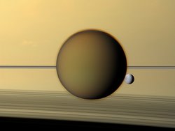 Titan vor den Ringen des Saturn. Um den Mond ist eine dünne, dunstige Hülle erkennbar. Halb verdeckt hinter Titan ist der erheblich kleinere Mond Dione zu erkennen.
