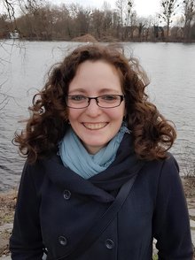 Porträt einer lächelnden Frau mit lockigem Haar, Brille, blauem Schal und dunklem Mantel vor einem Gewässer mit kahlen Bäumen im Hintergrund.