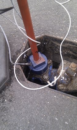 Ein Stahlrohr ist durch einen Gulli mit einer Wasserleitung verbunden. An dem Rohr sind mehrere weiße Kabel angebracht.